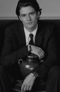 a man in a suit and tie sitting on a chair with a vase behind him