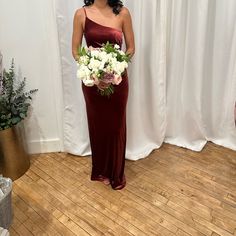 a woman standing in front of a white curtain holding a bouquet of flowers and wearing a burgundy dress