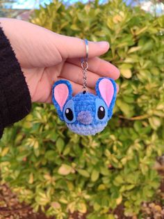 a hand holding a small stuffed animal keychain in front of some bushes and shrubbery