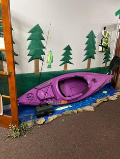 a purple kayak sitting on the ground next to a wall with trees painted on it