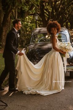 a woman in a wedding dress standing next to a man near an old fashioned car