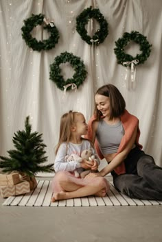 a mother and daughter sitting on the floor with presents in front of christmas wreaths