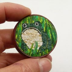 a hand holding a small button with a frog on it's face and grass in the background