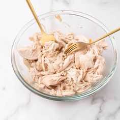 a glass bowl filled with shredded chicken and two gold forks on top of a marble counter