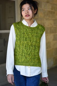 a woman wearing a green sweater and blue jeans holding a flower in her hand while standing next to a building