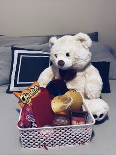 a white teddy bear sitting on top of a bed next to a basket filled with snacks