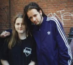 two people standing next to each other in front of a brick wall with graffiti on it