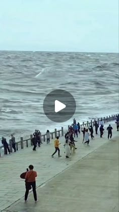 people are walking on the pier near the water and waves in the ocean, while one person is holding a surfboard