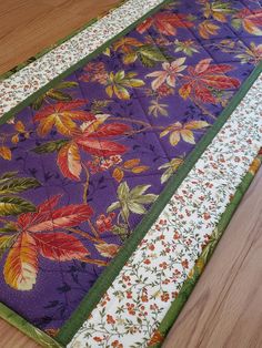 a quilted table runner with flowers and leaves on purple fabric, sitting on a wooden floor