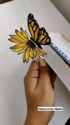 a hand holding a yellow and black butterfly on top of a white piece of paper