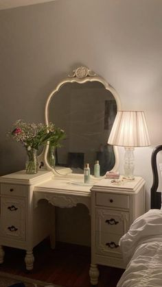 a white desk with a mirror, lamp and flowers on it next to a bed