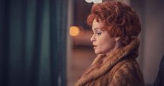 a woman with red hair wearing a fur coat looking off into the distance in a dimly lit room