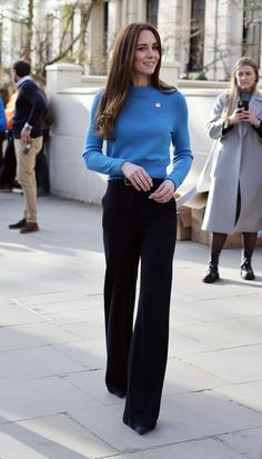 a woman in blue sweater and black pants standing on sidewalk with other people behind her