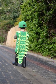 a little boy dressed up as a green dragon walking down the street with his hat on