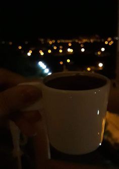 a person holding a coffee cup in their hand at night with city lights in the background