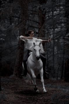 a woman riding on the back of a white horse through a forest filled with trees