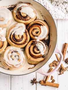 cinnamon rolls with icing in a skillet on a white wooden table next to cinnamon sticks