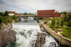 a river running through a lush green park next to a tall red brick building with a bridge over it