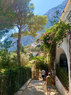 two people walking down a narrow street with trees and bushes on either side of the road