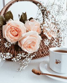 a basket filled with pink roses next to a cup of coffee and saucer on a table