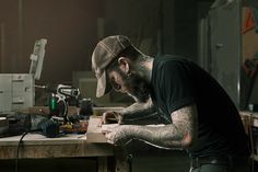 a man with tattoos working on a piece of wood at a table in a workshop