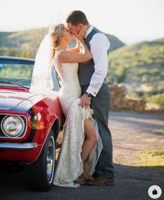 a bride and groom kissing in front of a red car on the side of the road