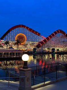 an amusement park at night with lights on the water and roller coasters in the background