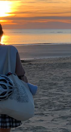 a woman walking on the beach at sunset with her bike helmet in hand and bag over her shoulder