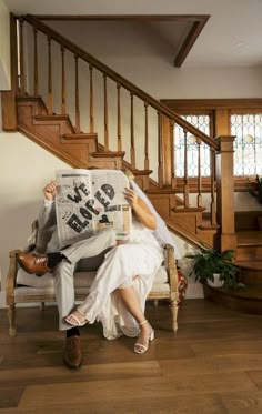 a bride and groom sitting on a couch reading the newspaper in front of their staircase