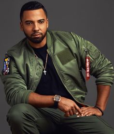 a man in a green jacket and black t - shirt is sitting on a stool