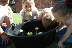 three girls and one boy are playing with apples in a large black tub on the grass