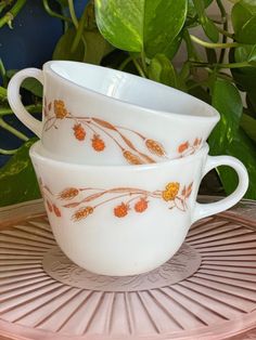 two white cups sitting on top of a pink tray next to green leaves and plants