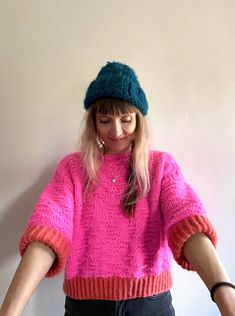 a woman wearing a pink sweater and green hat with her hands in the air while standing next to a white wall