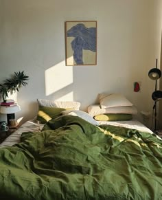 an unmade bed with green sheets and pillows in a white room next to a potted plant