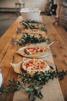 several pizzas on plates lined up on a long table with greenery around them