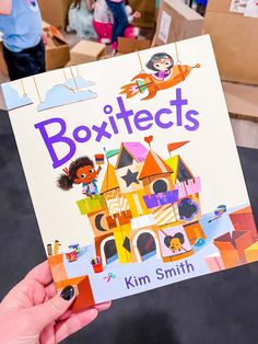 a person holding up a children's book in their hand with boxes behind them