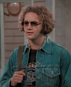 a man with curly hair and sunglasses standing in front of a house wearing a jean jacket