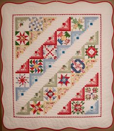 a white quilt with red, green and blue designs on the border is hanging in front of a brown wall