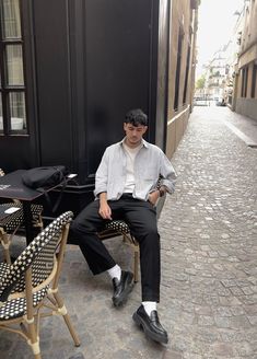 Boy sitting on a Parisian street chair in Le Marais, wearing a striped button-down shirt, pleated trousers from Uniqlo, and black shoes, against a stone street backdrop. Men Loafers Outfit, Loafer Outfits, Black Loafers Men, Black Trousers Men, Striped Shirt Men, Parisian Street, Loafers Outfit