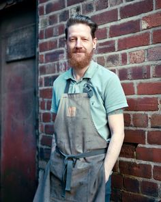 a bearded man wearing an apron standing in front of a brick wall with his hands on his hips