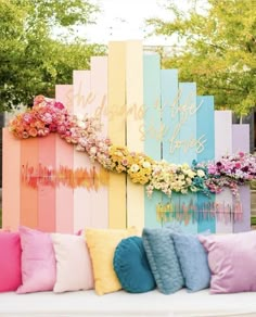 a bench with colorful pillows and flowers on it in front of a wooden fence that says,