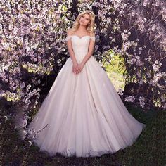 a woman standing in front of purple flowers wearing a wedding dress with off the shoulder sleeves