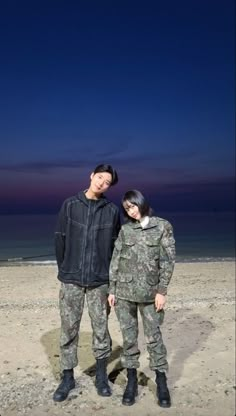 two people standing next to each other on a sandy beach at night with the sky in the background