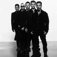 four men standing in front of a white wall wearing black clothing and sunglasses, all looking at the camera