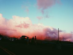 two people are standing in front of some cars on the side of the road with clouds behind them