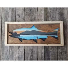 a wooden wall hanging with an image of a fish on it's side and mountains in the background