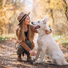 a woman kneeling down next to a white dog on a forest path with trees in the background