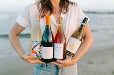 a woman holding three bottles of wine in front of her body and the ocean behind her