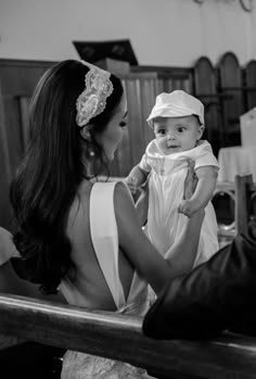 a woman holding a baby wearing a white dress and headband while sitting on a bench