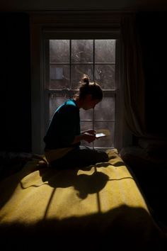 a person sitting on a bed reading a book in front of a window at night